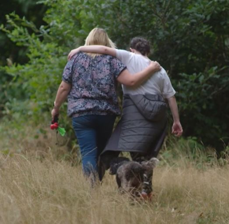 A woman with blonde hair wearing a blue patterned top walks through long grass with a girl with dark hair, their arms around each other's shoulders. A small grey dog walks behind them.