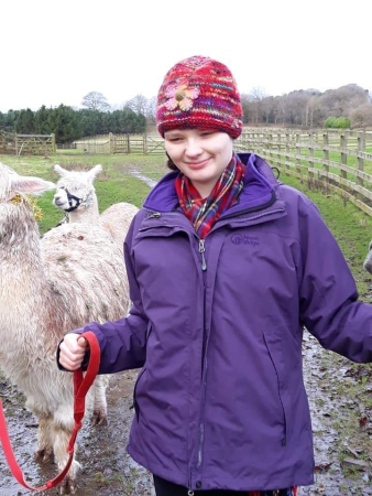 A girl in a purple coat and a pink hat with a flower holds the reins of a llama in a field.