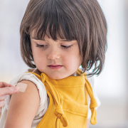 A female doctor in blue scrubs puts a plaster on the arm of a small girl wearing yellow dungarees, who looks down at the plaster.