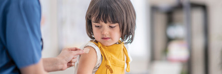 A female doctor in blue scrubs puts a plaster on the arm of a small girl wearing yellow dungarees, who looks down at the plaster.