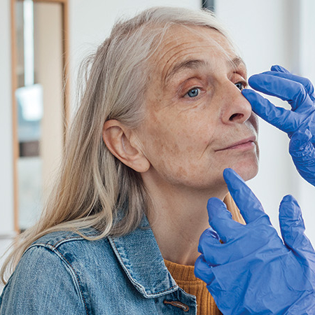 A female doctor is focused on a female patient's face as she is visually examining her. The patient is sitting down while the doctor treats her. It is an over-the-shoulder shot of the doctor examining the patient. The doctor is wearing a plastic apron and blue rubber gloves.