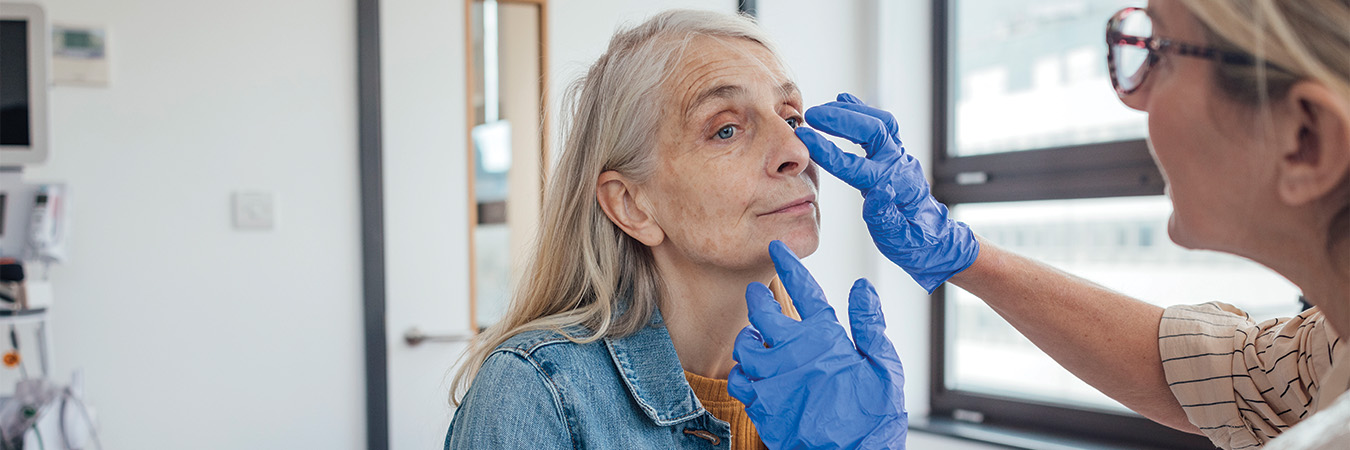 A female doctor is focused on a female patient's face as she is visually examining her. The patient is sitting down while the doctor treats her. It is an over-the-shoulder shot of the doctor examining the patient. The doctor is wearing a plastic apron and blue rubber gloves.