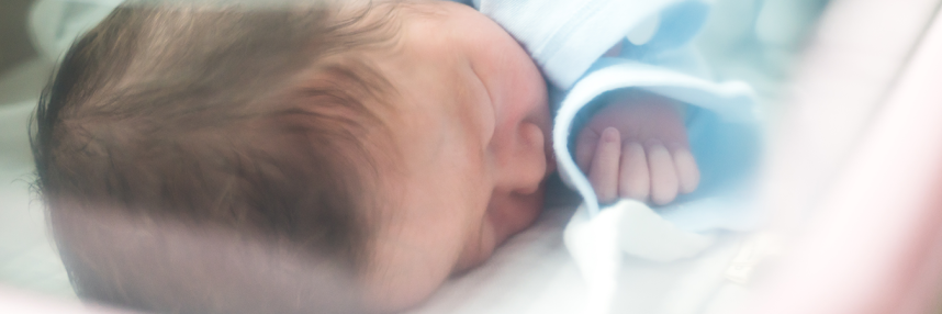 A baby in a blue sleep suit lies inside an incubator.