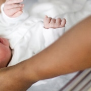 A newborn baby wearing a white bodysuit and hat lies in a cot, with an adult arm also in shot.