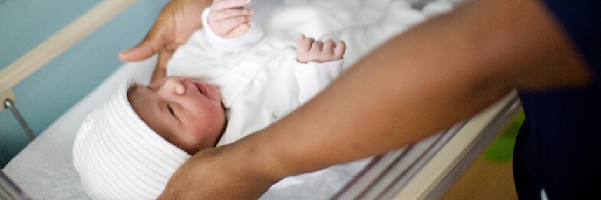 A newborn baby wearing a white bodysuit and hat lies in a cot, with an adult arm also in shot.