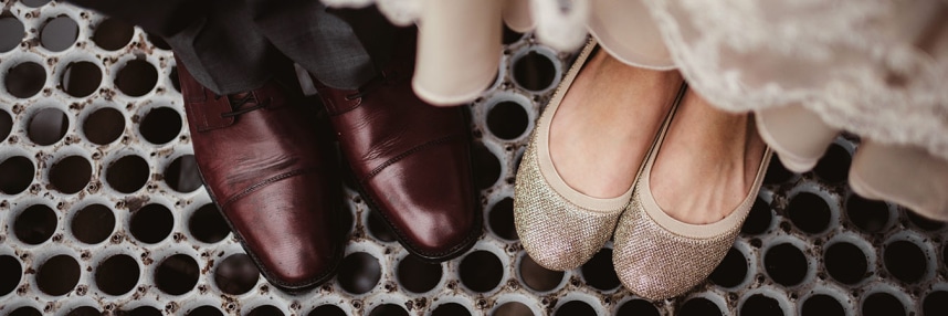 A close-up of a bride and groom's feet: the groom in shiny black shoes, the bride in white shoes. They stand on flooring with a geometric pattern.